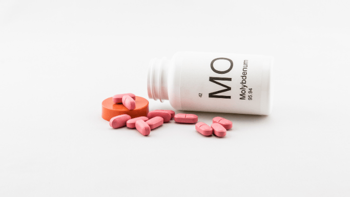 A white pill bottle labeled "Mo Molybdenum" is tipped over with several pink tablets spilled out onto a white surface.