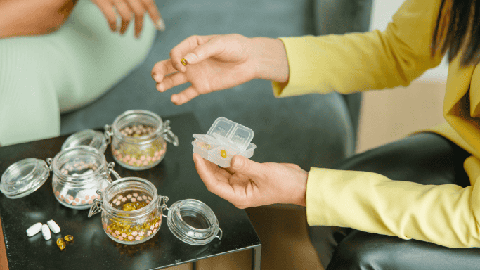 An image showing person holding a pill organizer with jars of pills on a table.