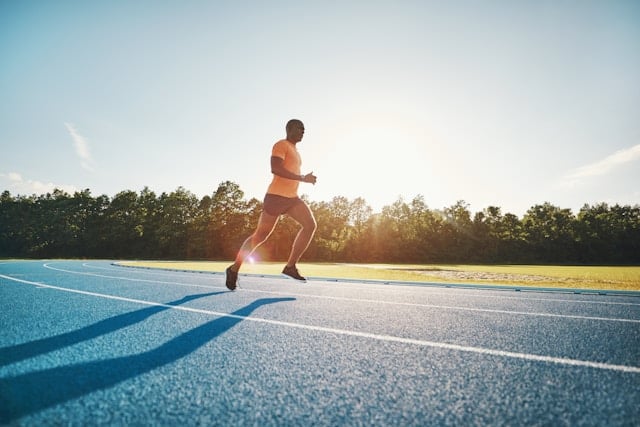 A runner sprints on a blue athletic track during golden hour with trees and sky visible.