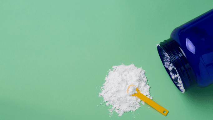 An open blue container of white powder, spilled onto a green surface with a yellow scoop resting in the powder.