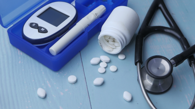 A diabetic kit with a glucose monitor, lancet, spilled white tablets, and a stethoscope on a blue wooden surface, representing conventional diabetes treatment.