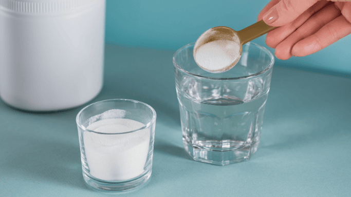 A hand is holding a gold spoon filled with white powdered supplement over a glass of water. 