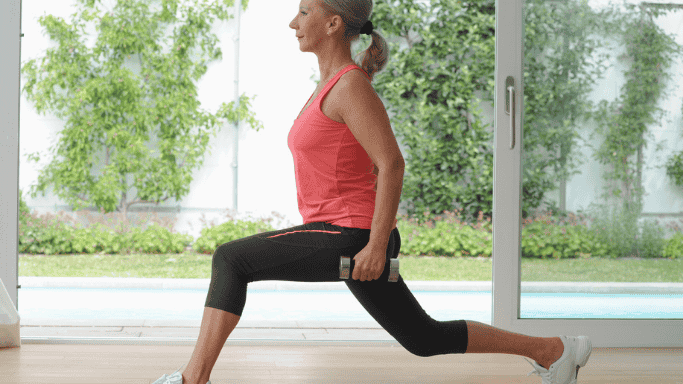 A fit woman performing a lunge exercise indoors while holding small dumbbells, wearing athletic clothes and standing in front of a large window with greenery outside.