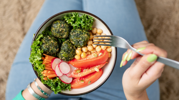 A bowl of colorful plant-based food including leafy greens, chickpeas, tomatoes, radishes, carrots, and spinach bites, held by a person with a fork.