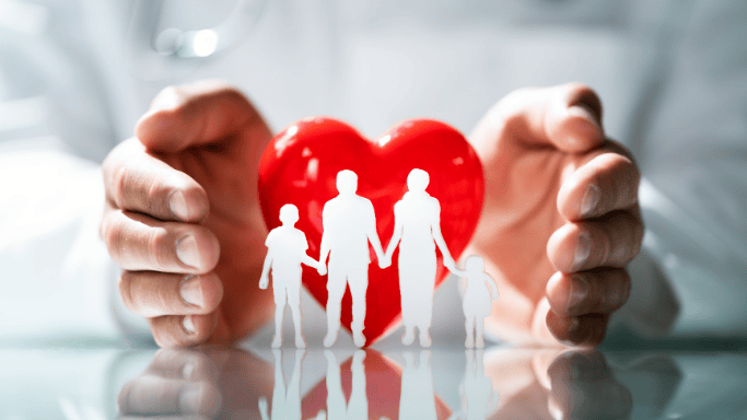 Hands protecting a red heart with a paper-cutout silhouette of a family, symbolizing healthcare, family protection, or insurance.