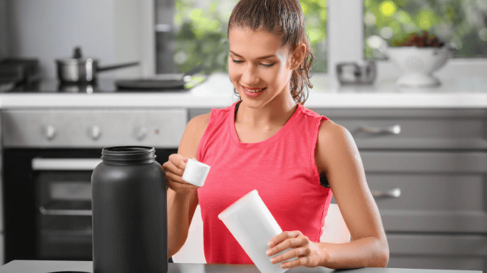 A woman in workout attire scooping protein powder into a shaker bottle from a large black container, suggesting supplement preparation post-workout.