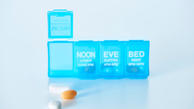 Blue pill organizer labeled noon, eve, and bed with several pills placed in front of it.