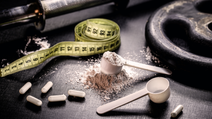 Supplement powder with scoops, capsules, a measuring tape, and gym equipment on a dark surface.
