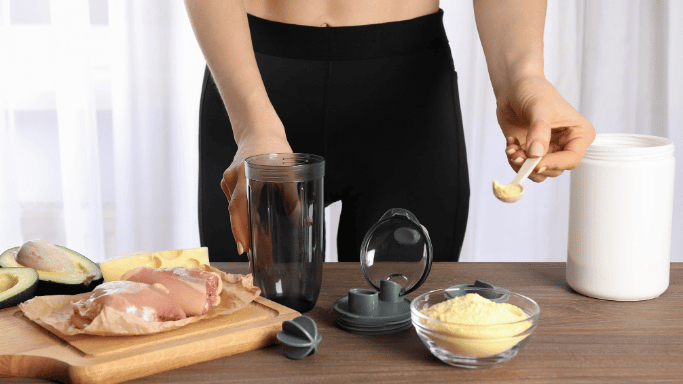 Person in black workout leggings preparing a protein shake, holding a shaker bottle and scooping protein powder, with raw chicken breast, avocado, and a bowl of powder on a wooden table.