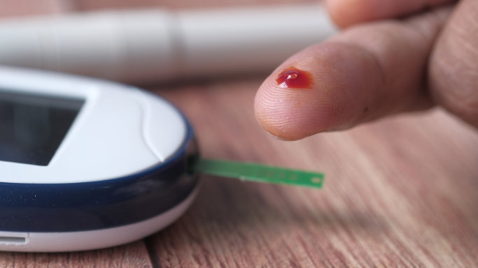 A close-up of a person’s fingertip with a drop of blood near a glucose meter and test strip, indicating a blood sugar test in progress.