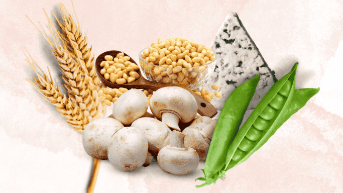 A collection of natural foods rich in spermidine, including wheat stalks, a wooden spoon and bowl filled with soybeans, a wedge of blue cheese, fresh green pea pods, and several white mushrooms arranged on a light background