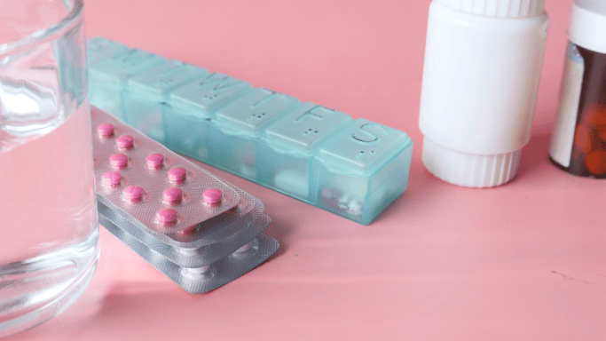 A collection of medication containers and pills on a pink surface. There are two blister packs with pink tablets, a blue weekly pill organizer, two white medicine bottles, a brown pill bottle, and a glass of water partially visible on the left side.