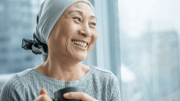hopeful woman wearing a headscarf, often associated with cancer treatment, smiles gently while holding a cup near a window. 
