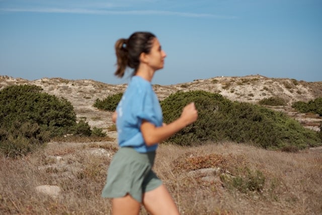 Woman in blue shirt and green shorts jogging through arid coastal landscape with low shrubs under clear blue sky.