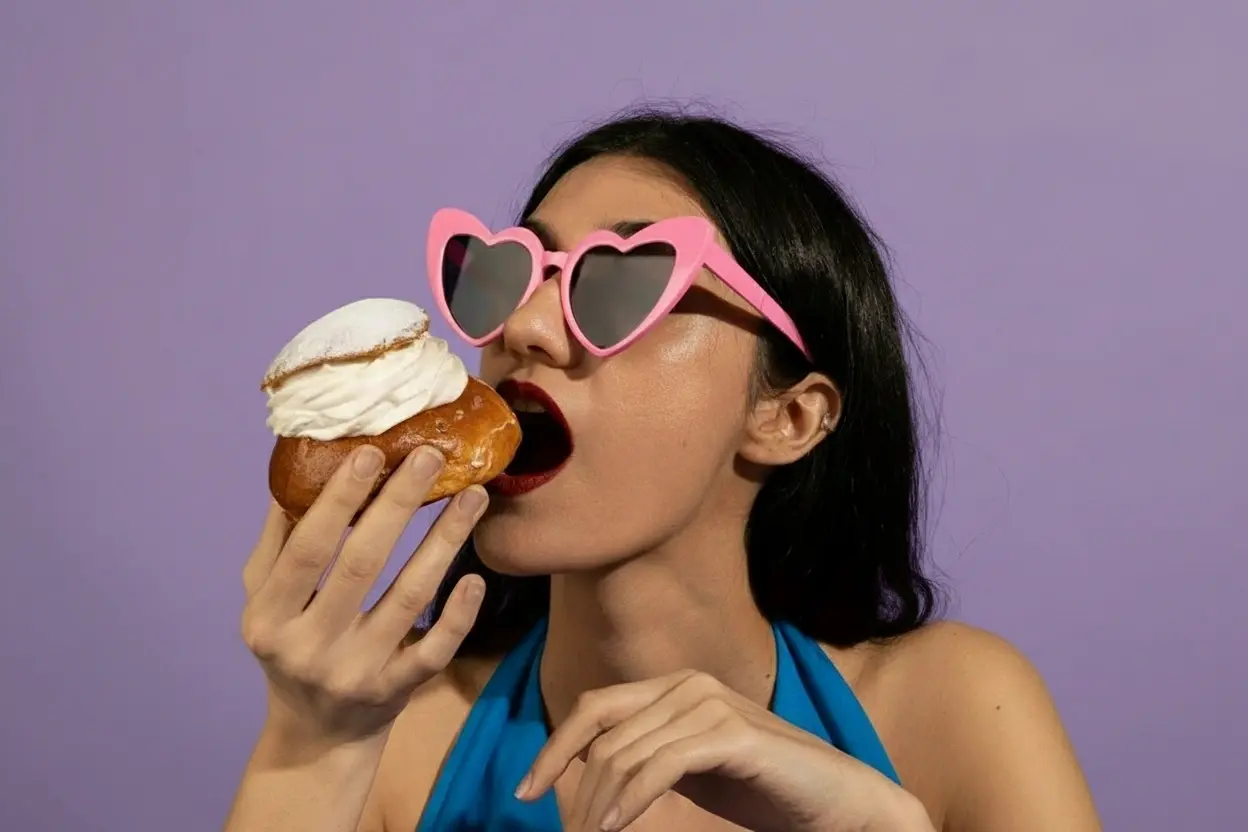 Woman wearing pink heart-shaped sunglasses takes bite of cream-filled pastry against vibrant purple background in playful portrait.