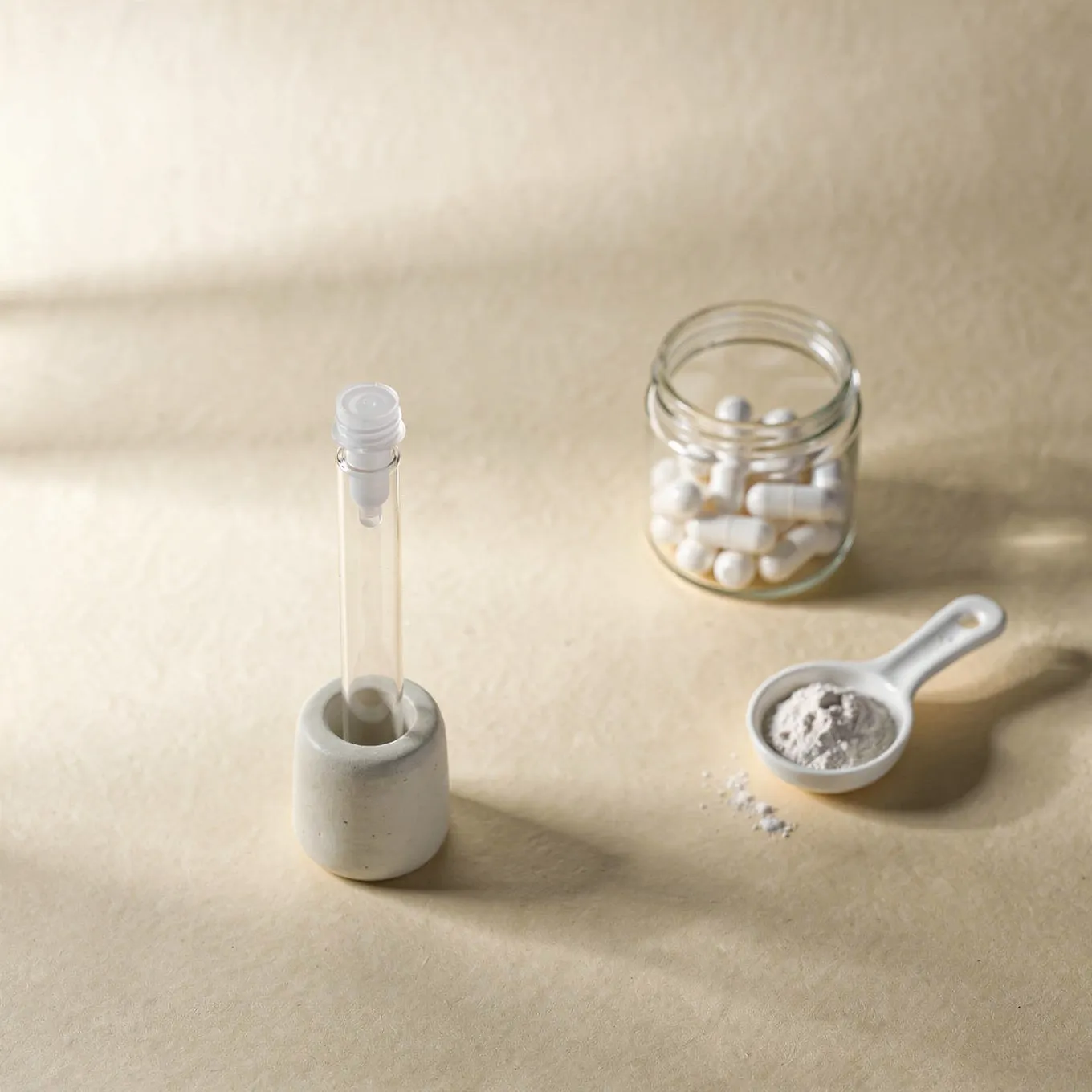 A sample tube in a white ceramic holder beside a glass jar of white capsules and a scoop of powder on cream paper