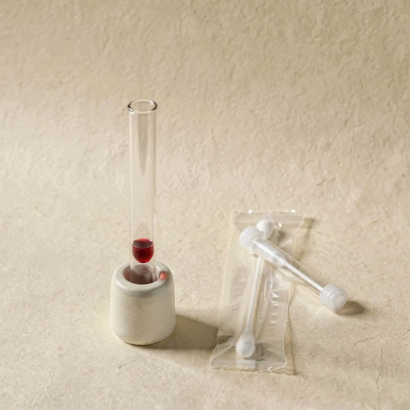 A sample tube in a white ceramic holder beside a sealed cheek swab in a clear tube on cream paper
