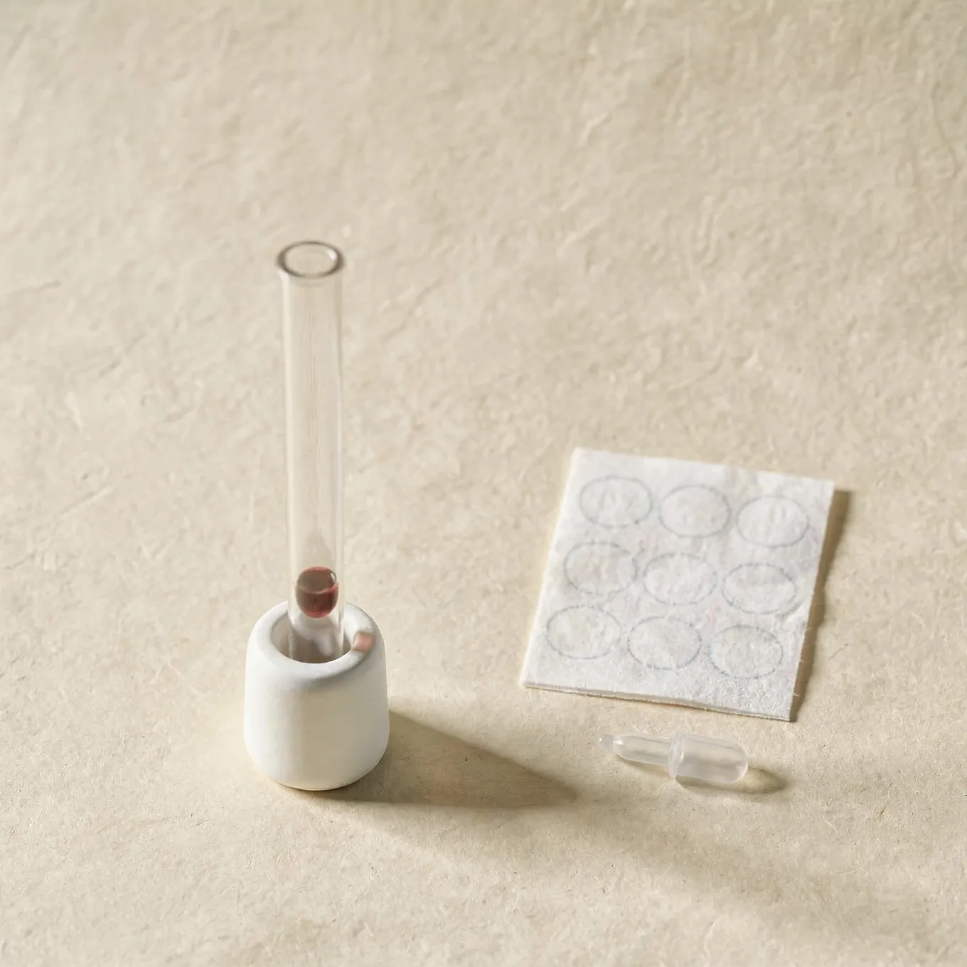 A sample tube in a white ceramic holder beside a blank blood spot card and a small lancet on cream paper
